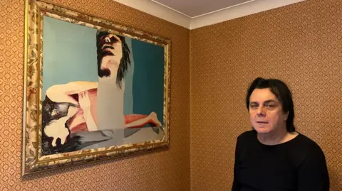 Martin Heath/BBC Michele Martignetti with long black hair wearing a black sweater and standing in front of a wall papered in orange. A modern framed painting showing a woman on a bed is hanging on the wall.