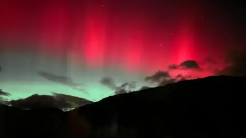 BBC Weather Watchers/Wee Von Bands of red aurora streak upward above a dark hillside, with green light near the horizon and scattered clouds drifting across the scene.