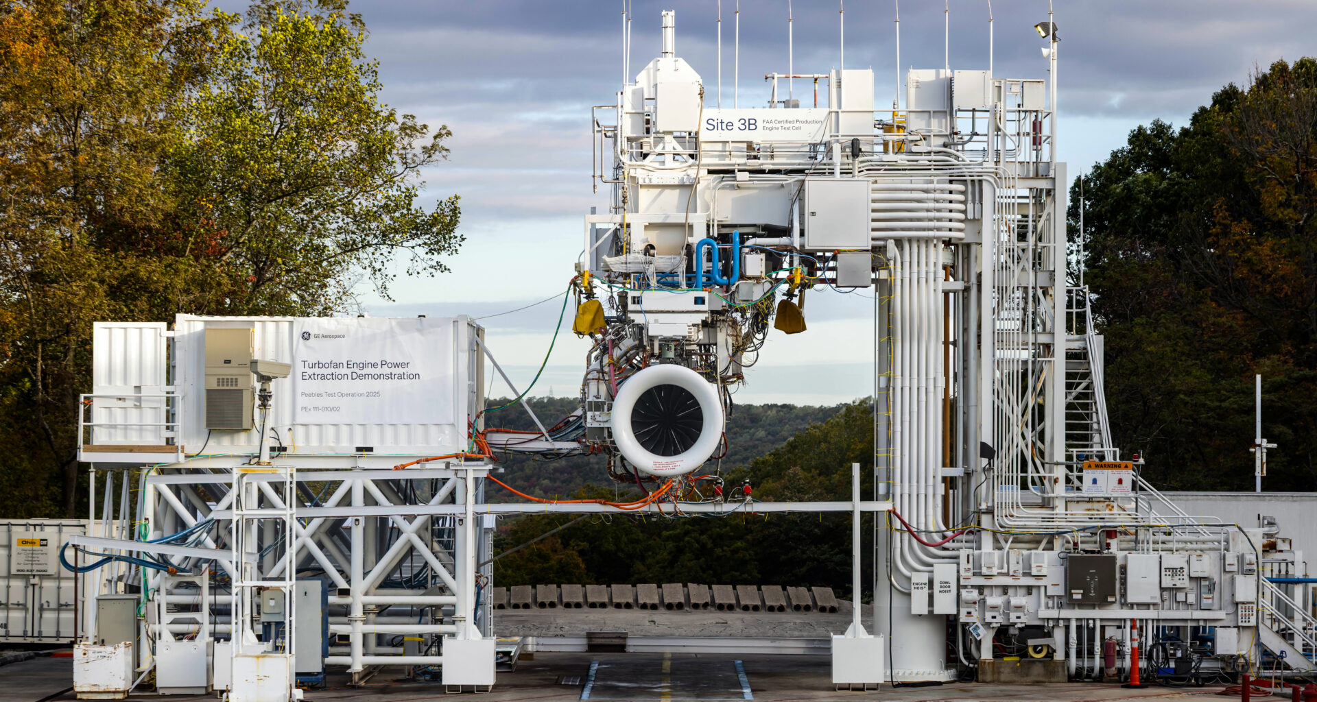 A hybrid engine, with its intake facing the camera, is mounted into a large, white scaffolding system. In the scaffolding structure to the hybrid engine’s left is a monitoring station in a modified shipping container. A series of pipes and wires to the engine’s right and above it, connecting the unit to the systems that provide it with fuel and collect data on its operations.