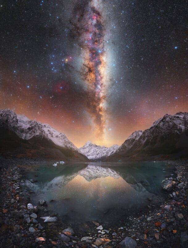 A star-filled night sky with the Milky Way visible above snowy mountains, reflected in a calm, rocky lake below; the scene glows with warm and cool tones, creating a dramatic, serene landscape.