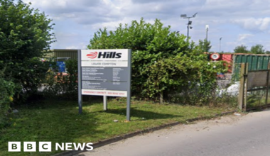 The entrance to a household recycling centre on a grey day. large metal fencing around it with huge red metal containers for recycling.
