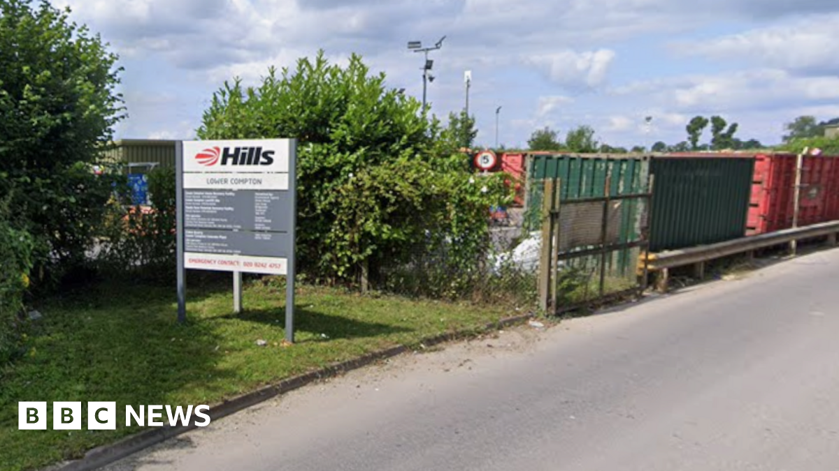 The entrance to a household recycling centre on a grey day. large metal fencing around it with huge red metal containers for recycling.