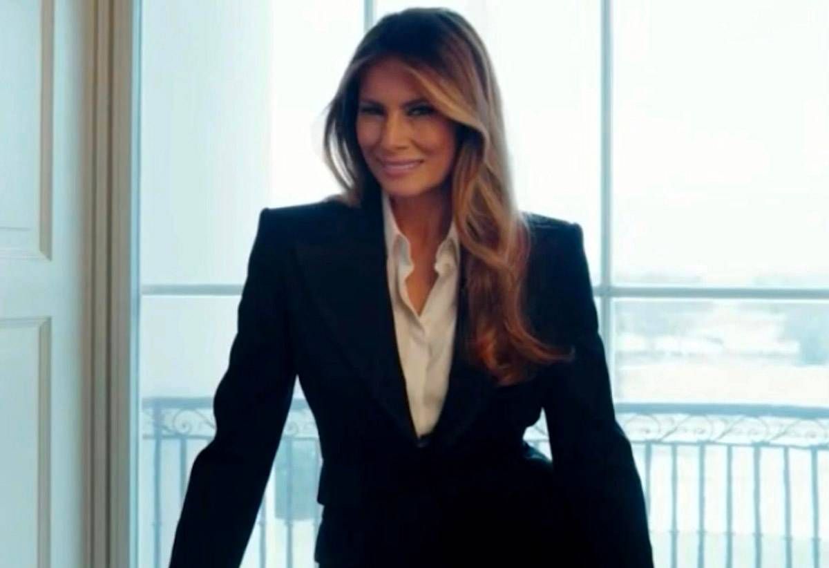 An individual in a tailored black blazer and white inner shirt stands confidently in a modern, well-lit room with large windows, showcasing an exterior view.