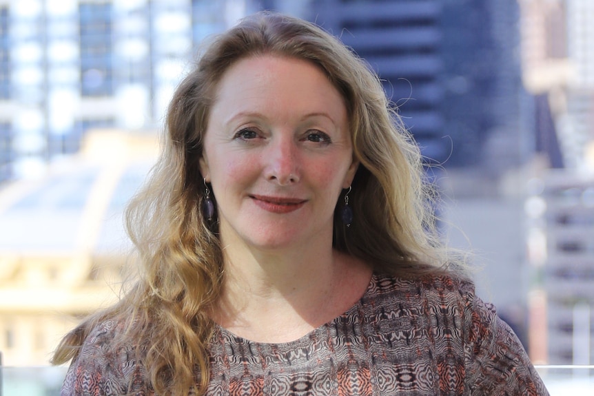 A middleaged blonde woman in a pattern top smiles for a portrait with a city in the background.