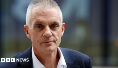 Helene Svinos sits on a sofa in between two dogs, with her arms around them. She is smiling and wearing a dark gilet over a patterned top, and leggings, with her dark hair tied back. There are cushions on the sofa and photo frames in the background.