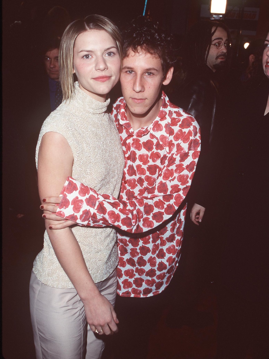 Man in floral shirt holds onto woman in cream dress. Both look at camera.