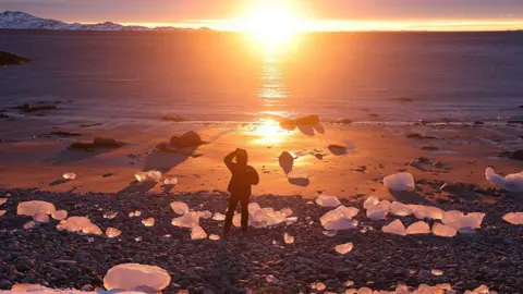 Getty Images A person stands on a beach at sunset among chunks of ice washed up on the shore in Nuuk, Greenland.