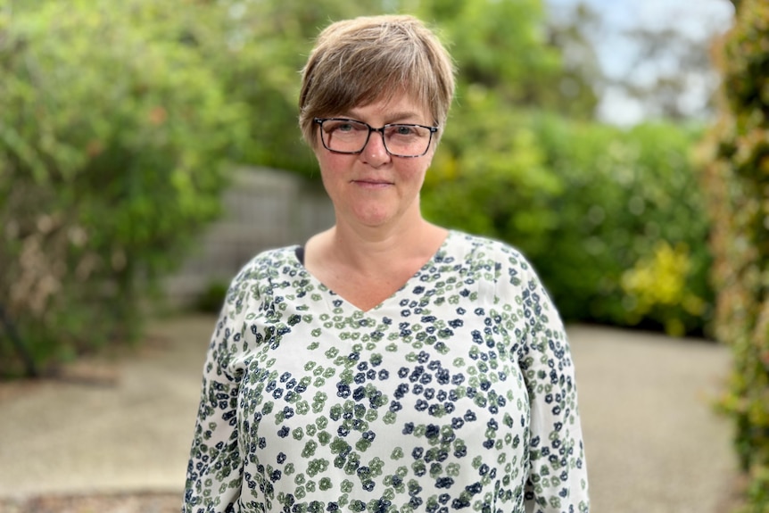 A middle-aged woman with short hair and glasses stands outdoors near some hedges.