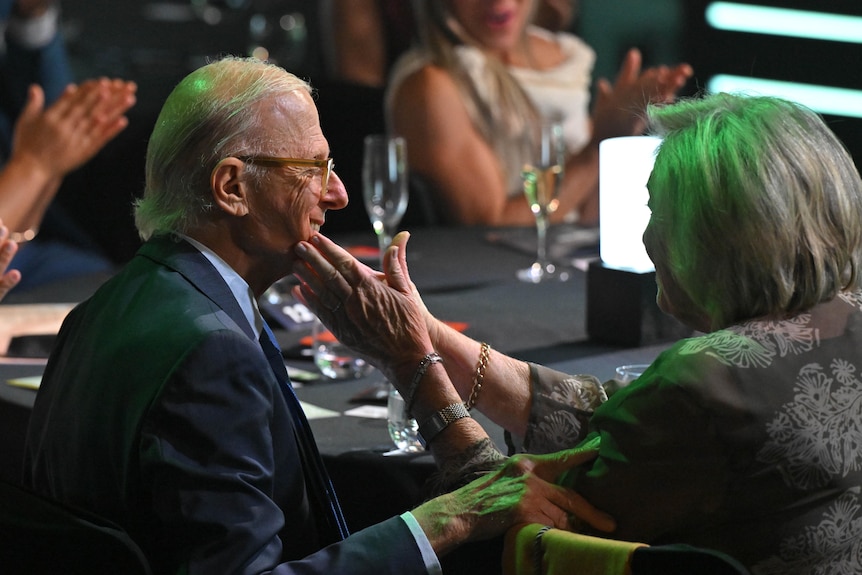 A woman holds her hands on the professor's face, at the awards, as people clap.