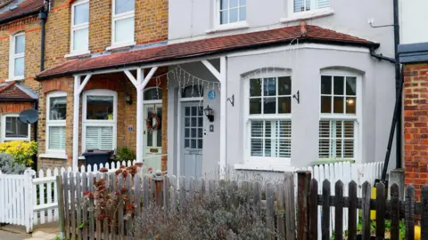 Heritage of London Trust A row of terraced houses with brick and painted facades, white-framed windows, small front gardens, and low wooden fences along a residential street.