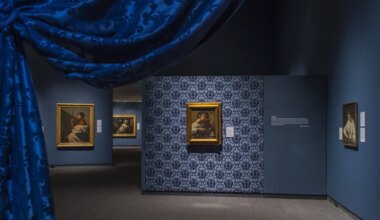 Interior view of an art gallery with framed paintings on blue patterned walls and a blue curtain in the foreground.