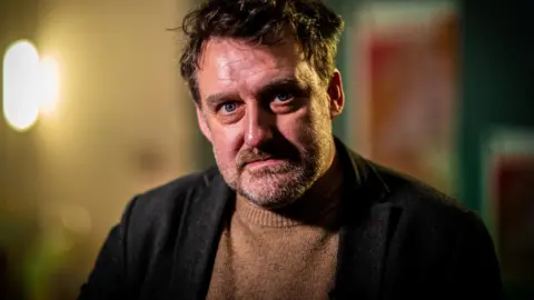 A man wearing a dark jacket over a light brown knit jumper, sitting indoors with warm lighting and soft, out‑of‑focus posters and décor in the background.