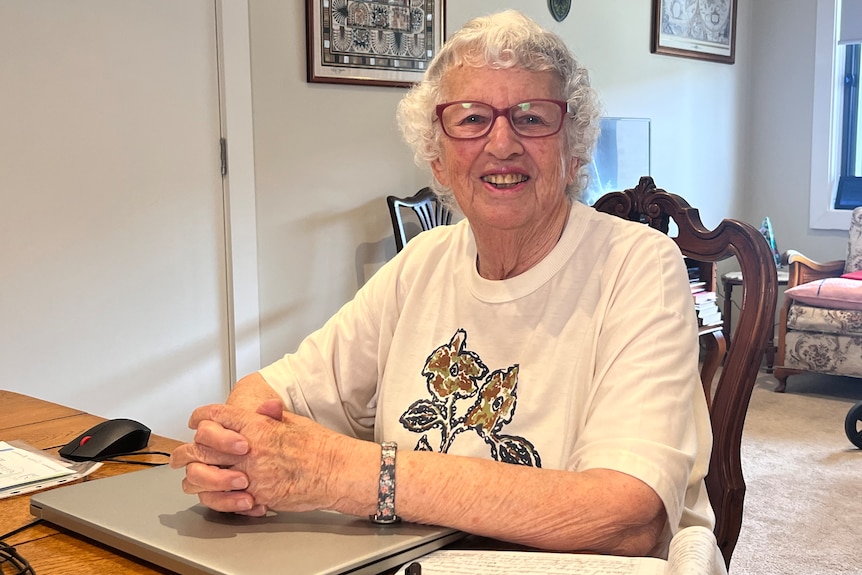 Older woman in a white t-shirt sitting at a desk with her hands on a closed laptop.
