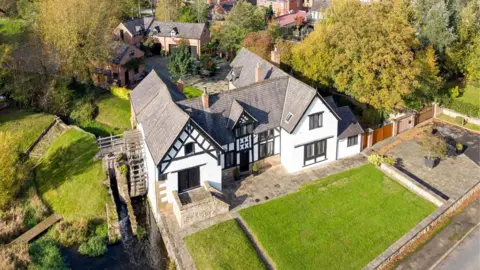 Matt Elson An aerial view of a historic white‑painted watermill and adjoining house with black timber framing, set beside a narrow river. A large wooden waterwheel sits on one side of the building. The property is surrounded by green lawns, stone terraces and mature trees in autumn colours. Behind the mill is a courtyard with brick outbuildings, and beyond that, a village of red‑brick houses and winding streets. The landscape stretches into rolling countryside under a bright sky with scattered clouds.