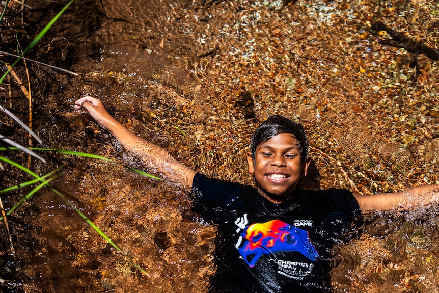 A young Indigenous boy lies on his back in clear water outside in the sun.