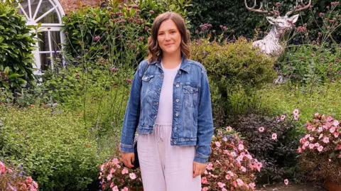 Sophie West A smiling young woman wearing a denim jacket poses for a photo in front of a colourful corner of a garden. Her shoulder-length brunette hair falls into loose curls at the sides of her head.