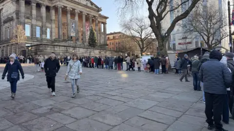 A large queue of people snakes out from the front of a Georgian period museum building with Palladian columns. 