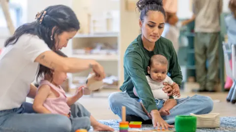 FatCamera/GettyImages Two women and two young children sitting on the floor in a playroom. One woman holds a baby while reaching towards a set of colourful toys on the rug. The other woman sits beside a toddler who is playing with wooden toys. Shelving with baskets and play materials is visible in the background.