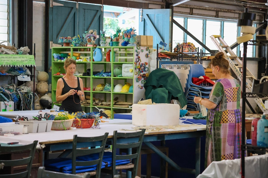 Two women standing at a table inside a shed hold pieces of string among shelves of colourful materials.
