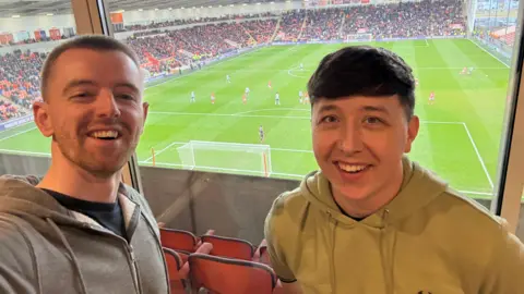 Dazza Darren Dowling (left) with short brown hair wearing a black t-shirt and grey hoodie and David Handling (right) with short brown hair and wearing a light green hooded top. They are standing in front of windows in a hotel room showing a pitch side view of Blackpool FC playing Bolton Wanderers with packed crowds. They are both smiling.