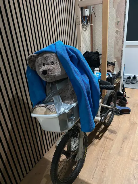 Michael Meighan Photograph of a large grey teddy bear attached to a washing basket at the front of a pedal bike. A blue blanket can be seen draped over the teddy's head. The bike is placed inside a kitchen area of a home, shoes can be seen behind it on the wooden floor.
