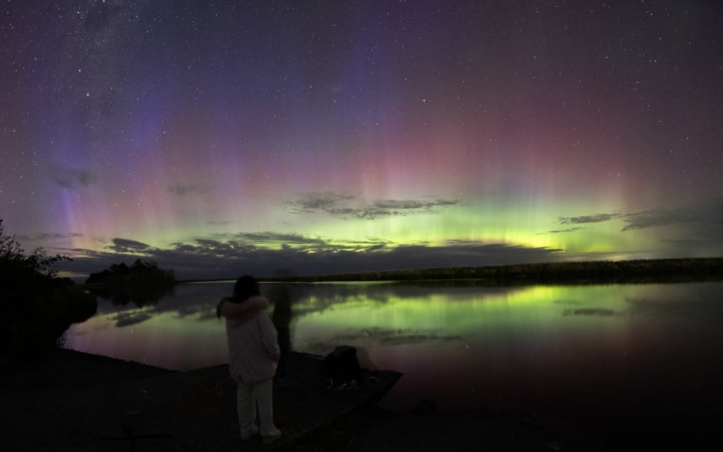 People look at the Aurora Australis, also known as the Southern Lights, as it glows on the horizon over the waters of Lake Ellesmere on the outskirts of Christchurch, New Zealand, on January 21, 2026. The Southern Lights are a natural phenomenon of vibrant lights. Solar activity, particularly a coronal mass ejection that sends charged particles and gas into space, causes it. It typically takes these particles three days to reach the magnetic field lines at the north and south poles before they enter the atmosphere. Upon entering, the energy and particles combine with atmospheric gases to create a spectrum of colored lights in the sky. (Photo by Sanka Vidanagama/NurPhoto) (Photo by SANKA VIDANAGAMA / NurPhoto via AFP)