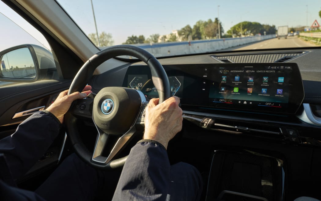 A driver's hand operates the steering wheel and reaches toward the curved digital infotainment screen of a BMW electric vehicle (EV) while driving on a street in Bari, Italy, on November 22, 2025. The wide screen displays the main menu, with icons for various apps and settings in Italian, such as 'Tutti' (All) and 'Infotainment', demonstrating the vehicle's integrated digital interface. (Photo by Matteo Della Torre/NurPhoto) (Photo by Matteo Della Torre / NurPhoto via AFP)