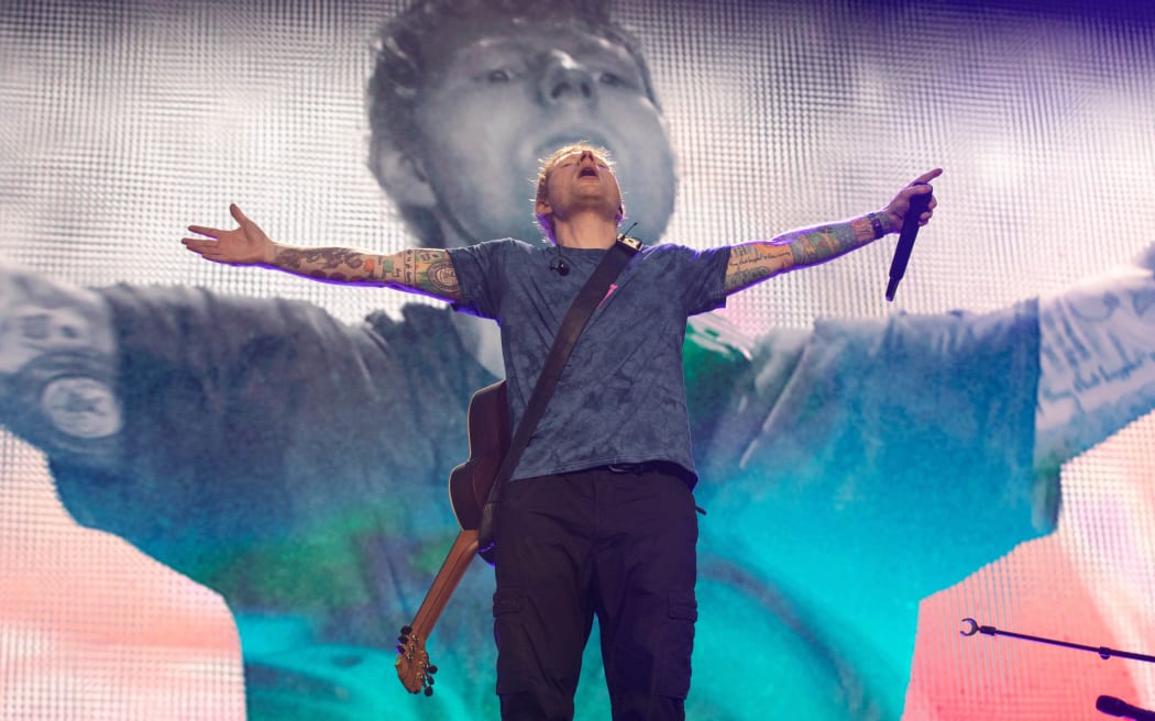 British pop star Ed Sheeran on stage with his arms wide open, mic in left hand and guitar slung on shoulder.