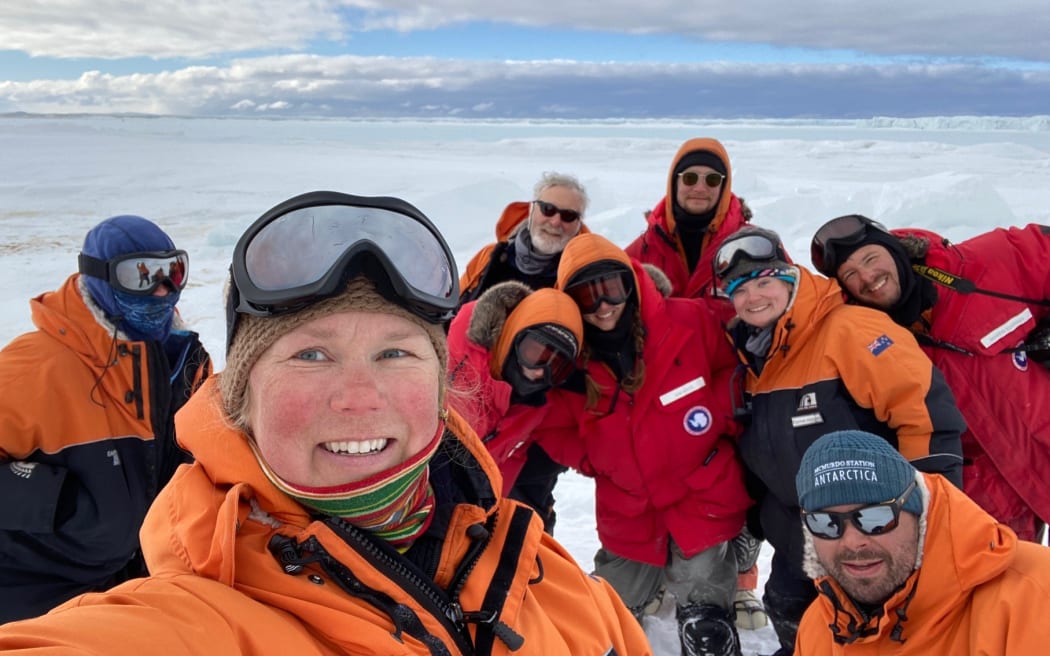 A wide-shot selfies of nine people on the ice beneath a cloudy sky. About half are wearing red jackets, and the others wearing black-and-orange Antarctica New Zealand jackets. Most are wearing beanies or have their hoods up, and a few have ski goggles or sunnies on.