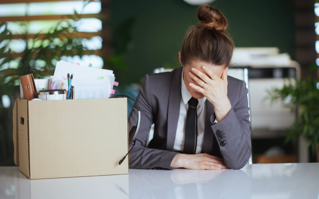 Stressed modern woman worker in modern green office in grey business suit with personal belongings in cardboard box.