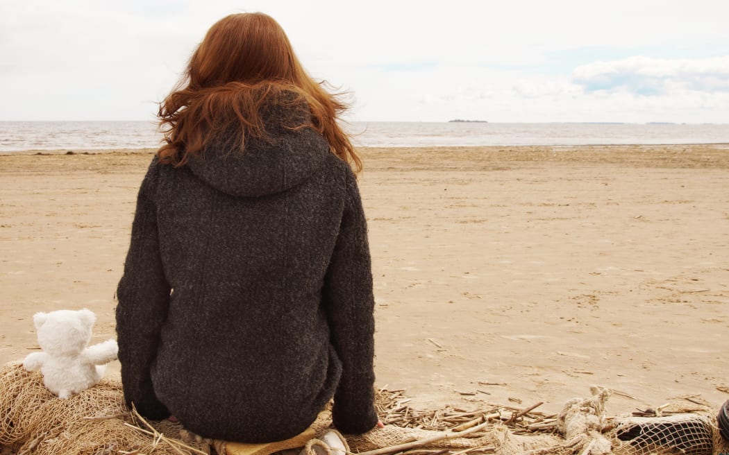 The girl sits on a fishing net and looks at the water of the bay, next to a teddy bear with her back.