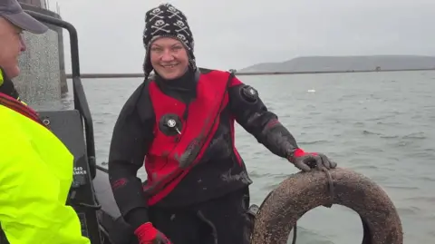The Ships Project Mallory Haas sitting on the edge of a boat holding a tyre which has been retrieved from the seabed in Plymouth Sound. She is wearing a black and red wetsuit and a black woolly hat with a white skull pattern. She is smiling widely. The sea and the sky are both quite grey.