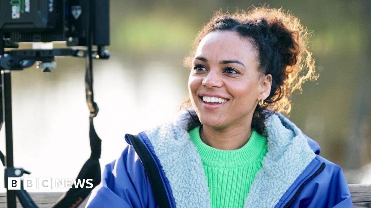 Cat White, a filmmaker, sitting down looking into a camera monitor. She is wearing a bright green jumper with a blue fleece-lined coat over the top. She is smiling but not looking at the camera.