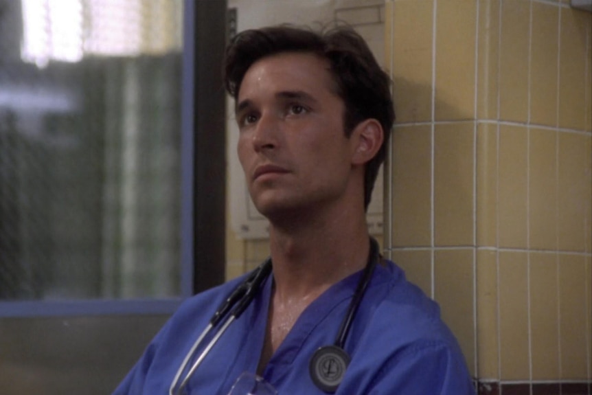 A TV still of Noah Wyle, 25 in 1996, looking contemplative in blue scrubs, stethoscope around his neck.