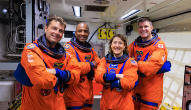 Four astronauts wearing an orange and blue spacesuit pose with their arms crossed inside of a facility.