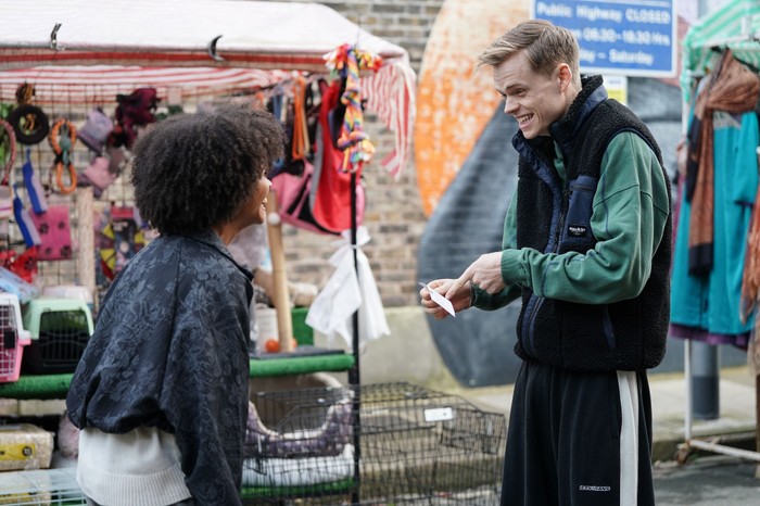 Oscar smiles, pointing at a piece of paper and looking at Jasmine in the market in a scene from EastEnders