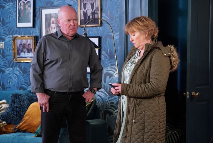 Phil stands looking at Julie, who is looking at her phone, in a scene from EastEnders