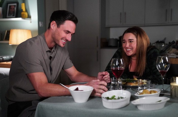 Vicki and Zack smile at a dining table surrounded by plates and a glass of wine in a scene from EastEnders