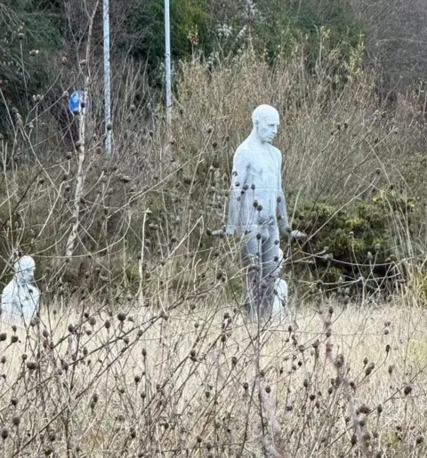 Holly Jones A pale, human-shaped sculpture stands among tall, dry weeds and grasses, partially obscured by vegetation in the foreground. Another similar sculpture is visible nearby, with trees and signposts in the background.