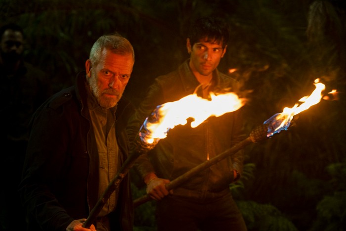 Hugh Laurie and Diego Calva star in The Night Manager season 2; in this scene, they are holding flaming torches and walking into a dark forest