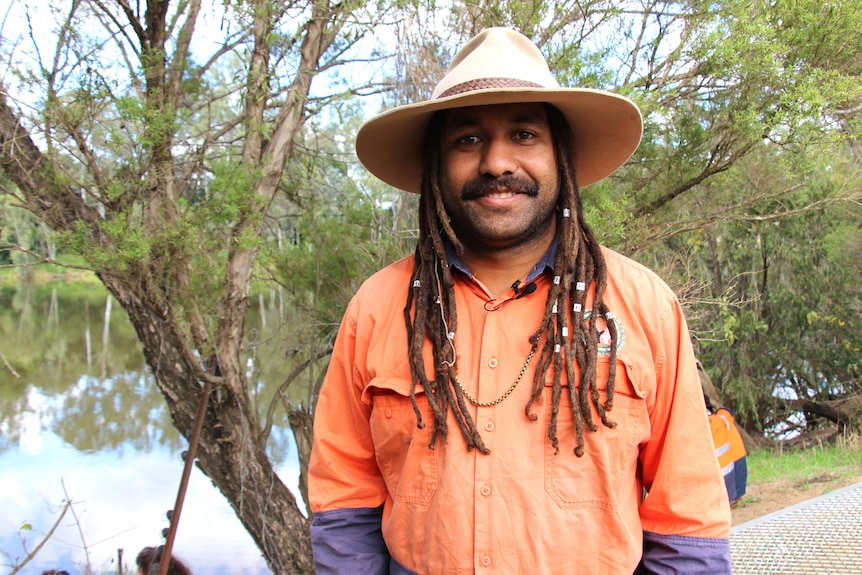 A man with dreadlocks wearing an orange shirt and hat with a river behind him.