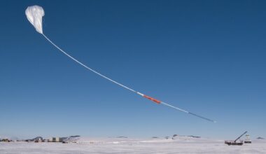 A white scientific balloon rises into the bright blue sky above snowy Antarctica. The sky and the balloon take up most of the image, with the ground only being a small white stripe at the bottom of the photo. The balloon has a long white "tail" with orange and black portions.