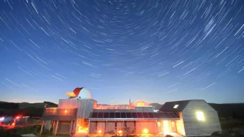 Marc Charron A brightly lit observatory building beneath a swirling starry sky