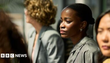 A young black woman with her hair tied back in a bun gazing with a concerned look on her face during a meeting with other female colleagues blurred out of focus around her