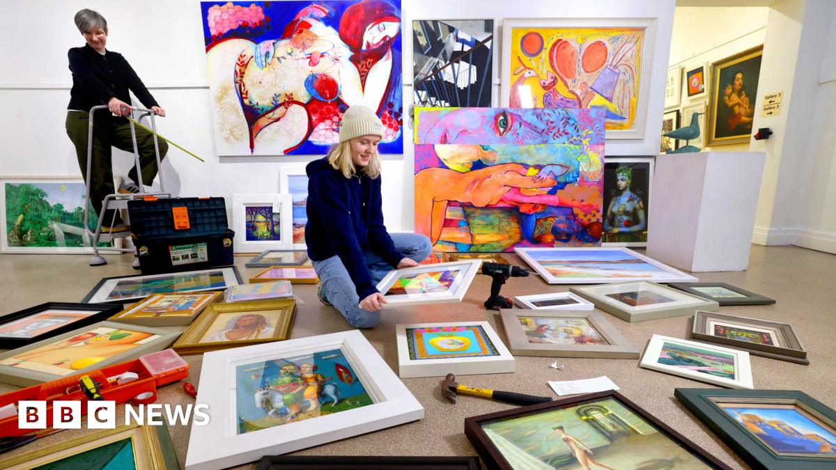 A woman sits on the floor of an art gallery, surrounded by paintings and artwork. Another woman is standing on a small stepladder in the background