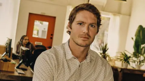 Yondr Graham Dugoni sits with his arms folded and looking directly at the camera. He is wearing a white shirt with black polka dots.