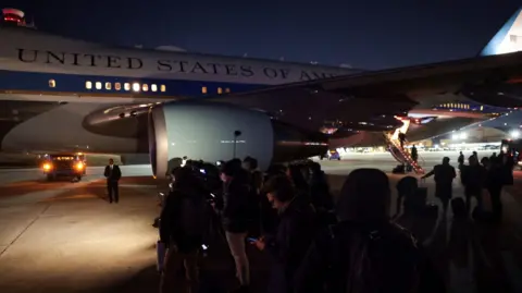 Reuters A group of journalists stand on the tarmac next to the Air Force One jet. The white and blue plane stands in the background, with the 'UNITED STATES OF AMERICA' logo visible on the side of the aircraft. The press wait in the dark, dressed in dark colours and checking their mobile phones.