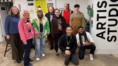 The Wilson A group of artists smile together inside a gallery space. They are wearing colourful clothes, including jumpers, hats and scarves. One of the signs on the wall says 'Artist Studio'. 