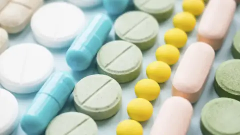 Getty Images A stock image of lines of pills from above - from left, a line of round white pills, a line of pale blue capsules, a line of pale green round pills, a line of bright yellow small round pills, a line of pink capsules, then a line of pale green round pills
