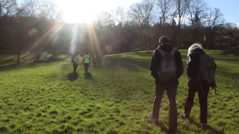 Bath Mind A group of people going for a walk in an open park on a sunny day with volunteers from Bath Mind. Trees and a line of houses can be seen in the distance.